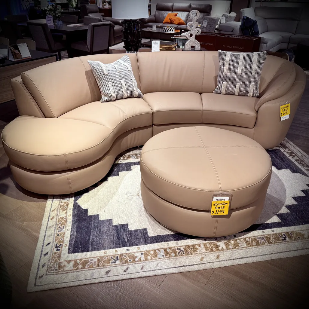 Curved beige leather sectional sofa with a matching round ottoman, styled with patterned throw pillows on a geometric area rug in a furniture showroom