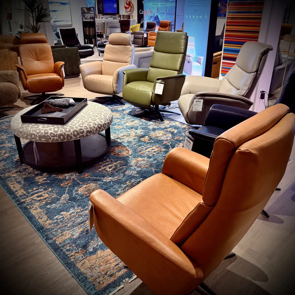 Group of leather recliner chairs in tan, green, and cream arranged around a round ottoman on a blue patterned rug in a showroom display